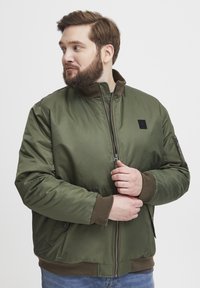 Maslinasto zeleni bomber jakna s visokim ovratnikom, zatvaranjem na zatvarač i rebrastim manžetama. Ima bočnu džepu i mali logotip na prsima.