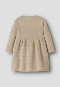 Beige neulemekko, jossa on pitkät hihat, teksturoitu päällinen ja liehuva pitsikuviollinen hame. Viimeistelty ribbaamalla kaula-aukkoa.