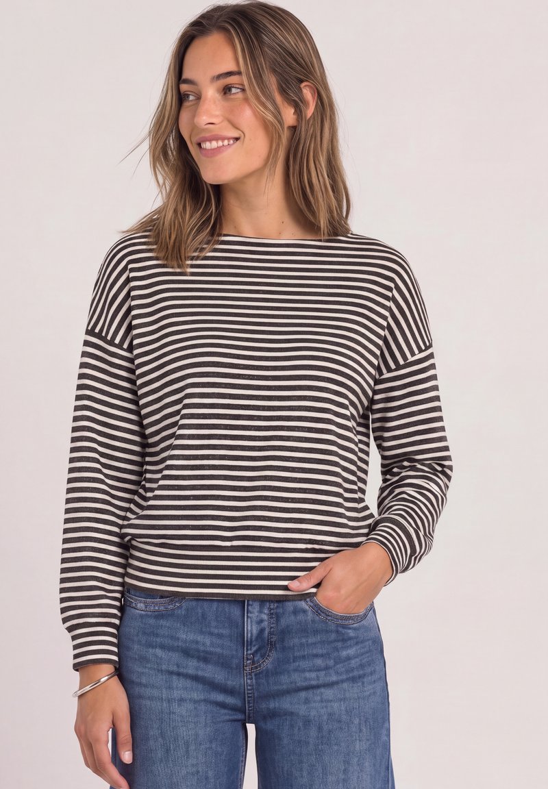 Femme aux cheveux mi-longs souriante, portant un haut à manches longues rayé noir et blanc et un jean bleu, la main dans la poche.