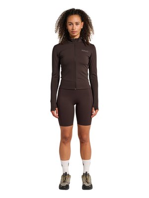 Kvinde stående med fronten mod kameraet iført en mørkebrun lynlåsjakke, matchende cykelshorts, hvide sokker og grå sneakers.