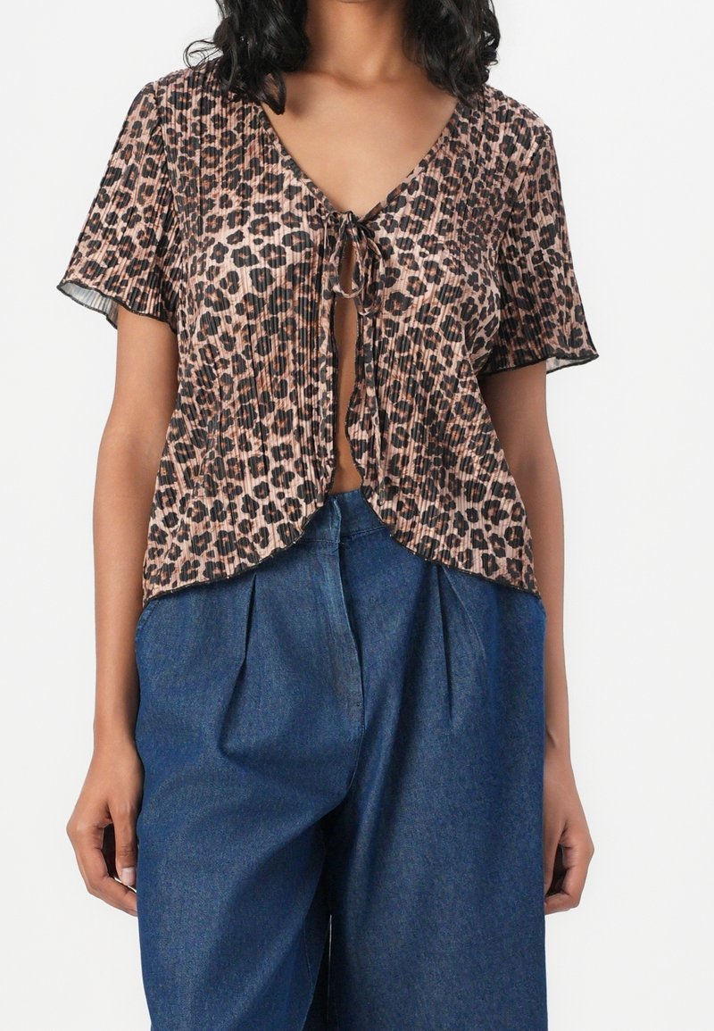 Femme portant un chemisier plissé à manches courtes imprimé léopard, noué à l'avant, associé à un pantalon bleu ample taille haute.