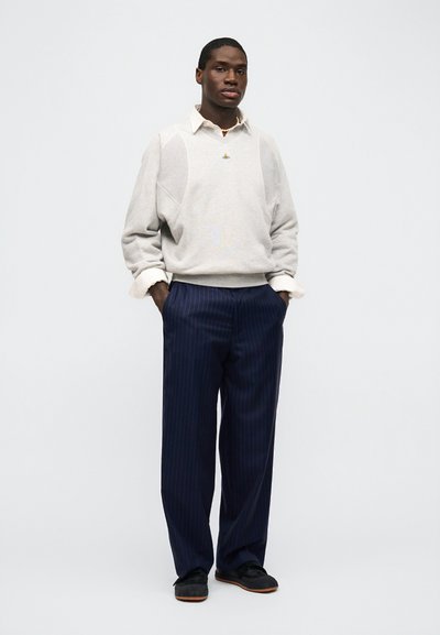 Άνδρας που στέκεται με τα χέρια στις τσέπες, φοράει λευκό πουκάμισο με γιακά κάτω από ανοιχτό γκρι πουλόβερ και φαρδιά παντελόνια με μπλε ναυτικό χρώμα και ρίγες.
