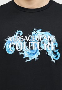 Μαύρο πουκάμισο με το κείμενο «Versace Jeans Couture» σε λευκό πάνω σε μπλε διακοσμητική πιτσιλιά και σχεδιασμό στροβίλου στο  στήθος.