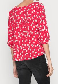 Blouse florale rouge avec des marguerites blanches, col rond, manches trois-quarts et détails froncés à l'arrière. Le tissu est léger avec une finition texturée.