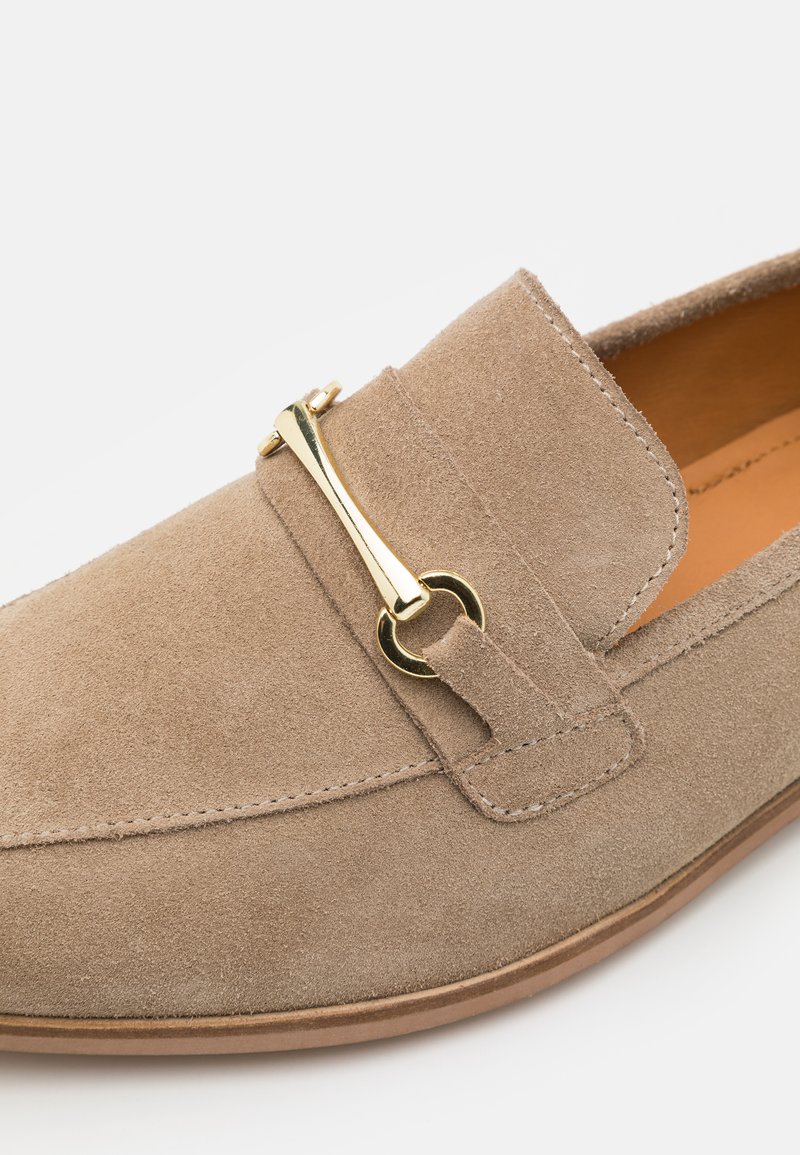 Mocassin en daim beige avec une texture lisse, présentant un accent en métal doré et des coutures visibles autour des bords supérieur et de la semelle.