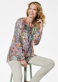 Mehrfarbige Langarmbluse mit floralen und geometrischen Mustern, Rundhalsauschnitt und gerafften Details an den Schultern. Kombiniert mit hellen Hosen.