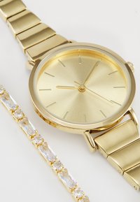 Montre dorée avec un cadran rond minimaliste et un bracelet en maillons métallique. Accompagnée d'un bracelet en or avec des pierres transparentes.