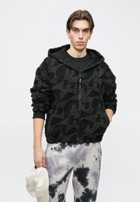 Melnā džemperī ar teksturētu rakstu, vilnas materiāls, rāvējslēdzēja aizdare un kapuce. Modelis valkā pelēkas kaklasaites krāsas bikses un tur baltu cepuri.