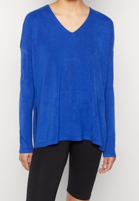 Femme portant un pull ample bleu à col en V avec des manches longues et un short noir ajusté longueur genou, debout devant un fond blanc.