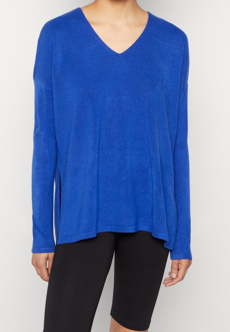 Femme portant un pull ample bleu à col en V avec des manches longues et un short noir ajusté longueur genou, debout devant un fond blanc.