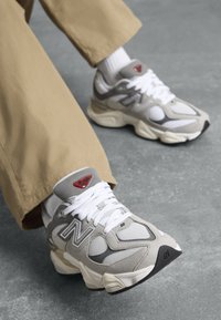En person iført beigefargede bukser og hvite sokker sitter, og viser grå og hvite New Balance-sneakers med kraftige såler på et grått gulv.