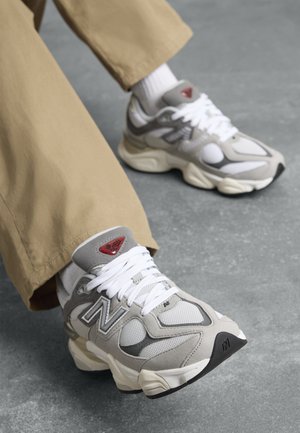 Person wearing beige pants and white socks seated, showing gray and white New Balance sneakers with chunky soles on a gray floor.