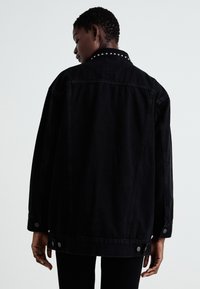 Black denim jacket featuring a studded collar, relaxed fit, and silver snap buttons on the cuffs and hem. Textured fabric with minimal detailing.