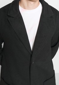 Persona che indossa un cappotto di lana nero sopra una maglietta bianca con collo tondo con lievi motivi tie-dye rosa, in piedi davanti a uno sfondo semplice.