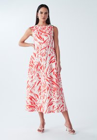 Kreisa krāsa maxi kleita ar sarkanu abstraktu rakstu, bez piedurknēm, teksturētu audumu un savilkumu pie kakla; kombinēta ar baltām sandālēm.