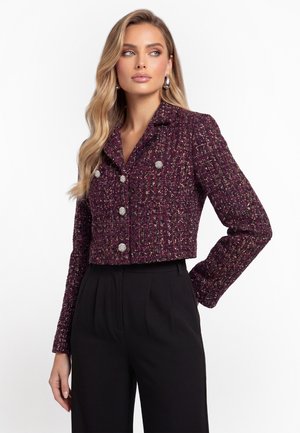 Donna che indossa un blazer corto, strutturato viola con bottoni d'argento e pantaloni neri a vita alta, in piedi con una mano sul fianco.