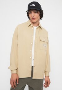 Giovane uomo che indossa una camicia beige con bottoni e una patch sul taschino, una maglietta bianca, pantaloni grigi e un cappellino nero di The North Face.
