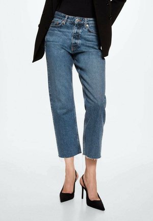 Femme portant un jean bleu court avec des ourlets bruts, un blazer noir et des chaussures slingback à talons hauts pointues noires, debout sur fond blanc.