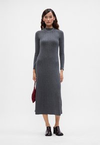 Siva rebrasta pletena obleka z ovratnikom za lažno turtleneck, dolgimi rokavi in ozkim krojem, v kombinaciji s sijajnimi črnimi čevlji in teksturirano rdečo torbico.