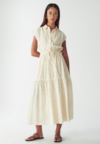 Krémové šaty s knoflíky a krátkým rukávem, elastickým pasem a volánkovou sukní. Mají černé puntíky a jsou vyrobeny z lehkého materiálu. Nošeny s černými sandály.