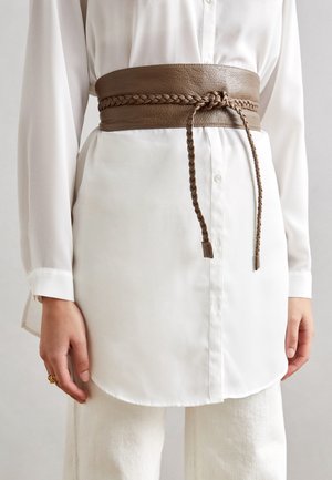 Brown wide leather belt with braided detail tied around waist over long white buttoned shirt and white pants.