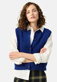 Blå stickad väst med diamondmönster, knappstängning fram och ribbad fåll, bärs över en vit skjorta med uppvikta ärmar. Rutig byxor synliga.