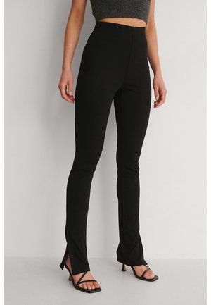 Frau trägt schwarze, hoch taillierte, enge Hosen mit vorderen Schlitzen kombiniert mit schwarzen Riemchen-Absatzsandalen und einem grauen Crop-Top.