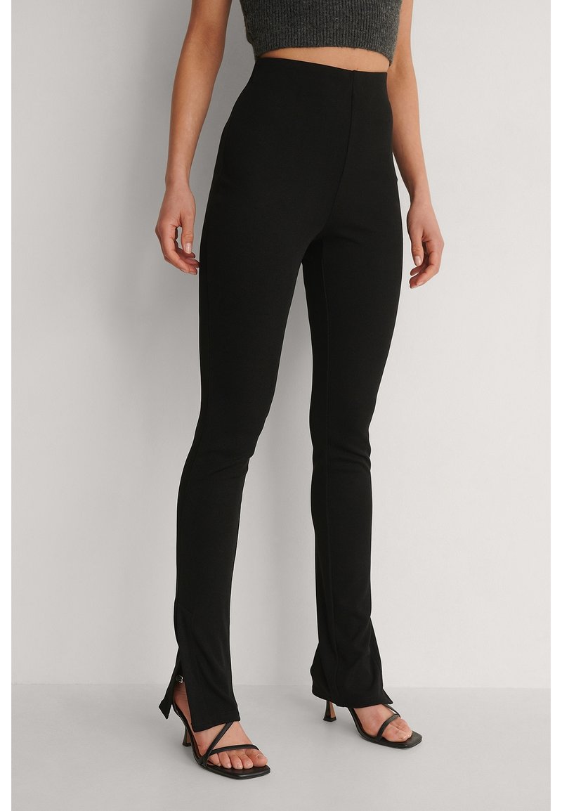 Femme portant un pantalon slim noir taille haute avec des fentes à l'avant, associé à des sandales à talons noires à brides et un crop top gris.