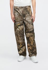 Καμουφλάζ παντελόνι σε καφέ και πράσινο χρώμα με σχέδιο φλοιού δέντρου. Διαθέτει ελαστική μέση και πλαϊνές τσέπες. Φοριέται με λευκά αθλητικά παπούτσια.