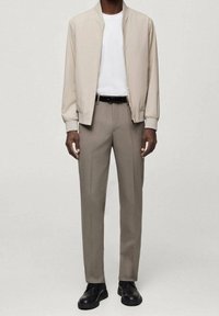 Beige Bomberjacke über einem weißen T-Shirt, kombiniert mit hellgrauen Hosen und schwarzen Schuhen. Die Jacke hat elastische Bündchen und einen Kragen.