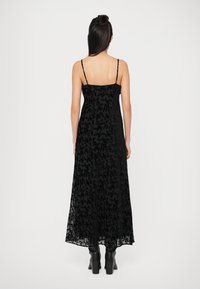 Abito lungo in pizzo floreale nero con spalline sottili, caratterizzato da una texture trasparente e una silhouette fluida. Indossato con stivali neri con tacco chunk.