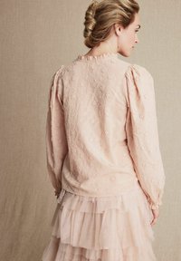Ljust rosa blus med blommönsterbroderi, puffärmar och volangkrage, tillsammans med en lager av tyllkjol. Mjuk, texturerad tyg.