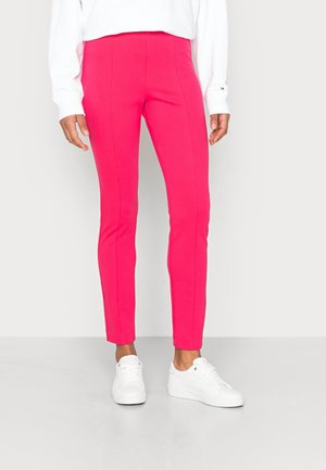 Persona con pantalones ajustados de color rosa brillante y zapatillas blancas, de pie contra un fondo claro y sencillo.