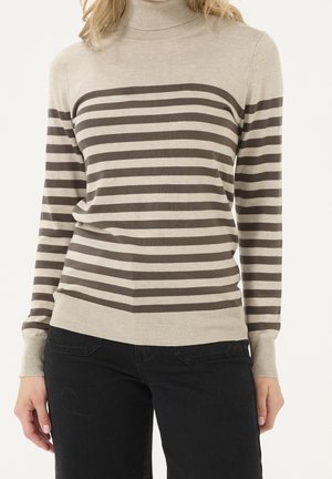 Nainen, joka pukeutunut beigeen pooloneuleeseen, jossa on tummanruskeita vaakaraitoja, ja mustiin housuihin, seisoo yksinkertaisen taustan edessä.