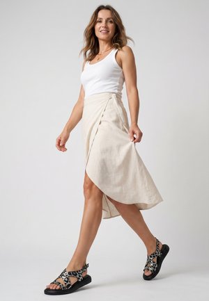 Frau geht, trägt ein weißes Tanktop, einen beigen Wickelrock und schwarz-weiß gepunktete Sandalen vor schlichten Hintergrund.