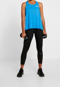 Blå atletisk linne med lös passform, med en logotyp. Svarta tajta leggings och svarta träningsskor. Håller hantlar i båda händerna.