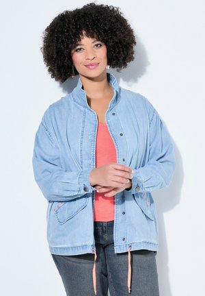 Frau mit lockigem Haar, die eine hellblaue Jeansjacke über einem korallenfarbenen Shirt und dunklen Jeans trägt, vor einem weißen Hintergrund stehend.