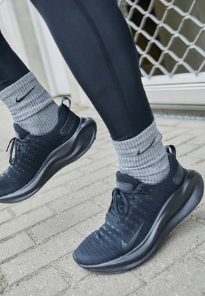 Des jambes dans des leggings noirs et des chaussettes grises Nike portant des chaussures de sport noires, s'élevant d'une surface pavée près d'une porte en métal.