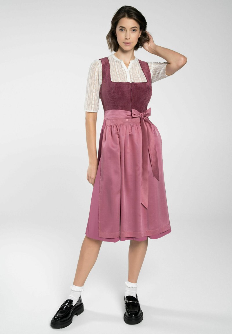 Rosa Kleid mit einem taillierten, strukturierten Oberteil und einem fließenden Satinenrock, der mit einer Schleife in der Taille gerafft ist. Weiße, gemusterte Bluse darunter.