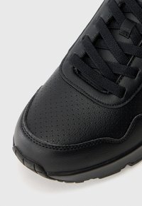 Chaussure de sport en cuir noir avec une texture perforée, un design à lacets et un embout lisse ; elle est dotée d'une semelle en caoutchouc noir et de détails de couture discrets.
