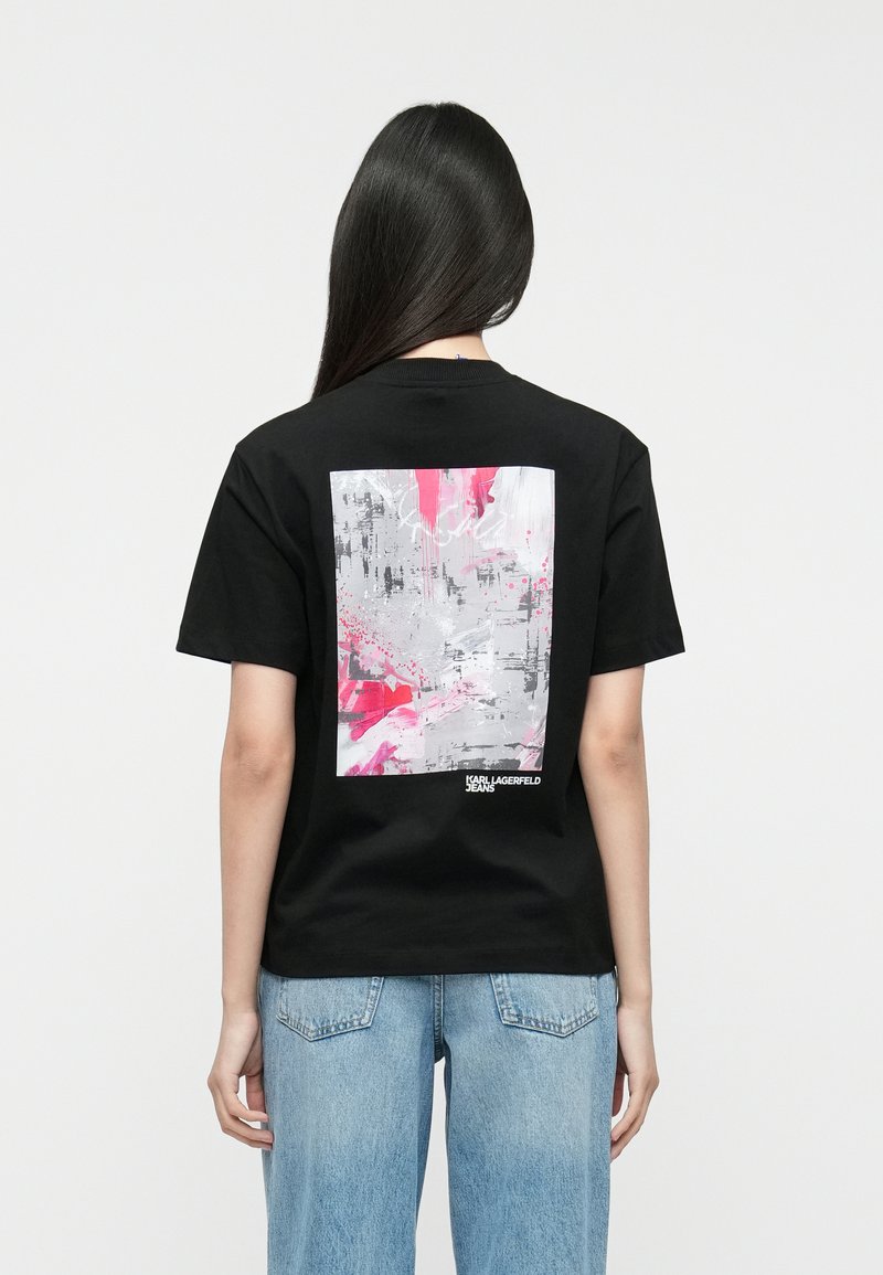 Čierne bavlnené tričko s veľkým abstraktným grafickým motívom v šedej a ružovej na zadnej strane, s textom "KARL LAGERFELD JEANS" na spodnej časti.