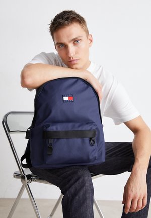 Młody mężczyzna z niebieskimi oczami siedzący na metalowym krześle, ubrany w białą koszulę i ciemne dżinsy, trzymający granatowy plecak Tommy Jeans.