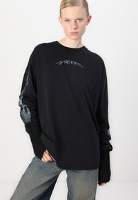 Crna majica dugih rukava s "RAGGED" logotipom u metalnom finišu na prsima, s teksturiranim crnim pletenim završecima i opuštenim krojem.