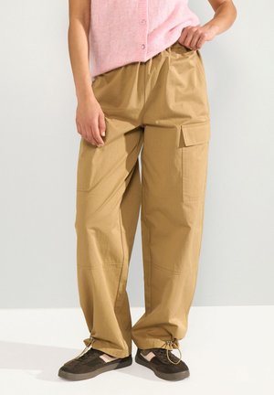 Persoon draagt losse beige cargobroek met trekkoorden aan de zoom, zwarte sneakers en een roze mouwloos topje, staand tegen een effen muur.
