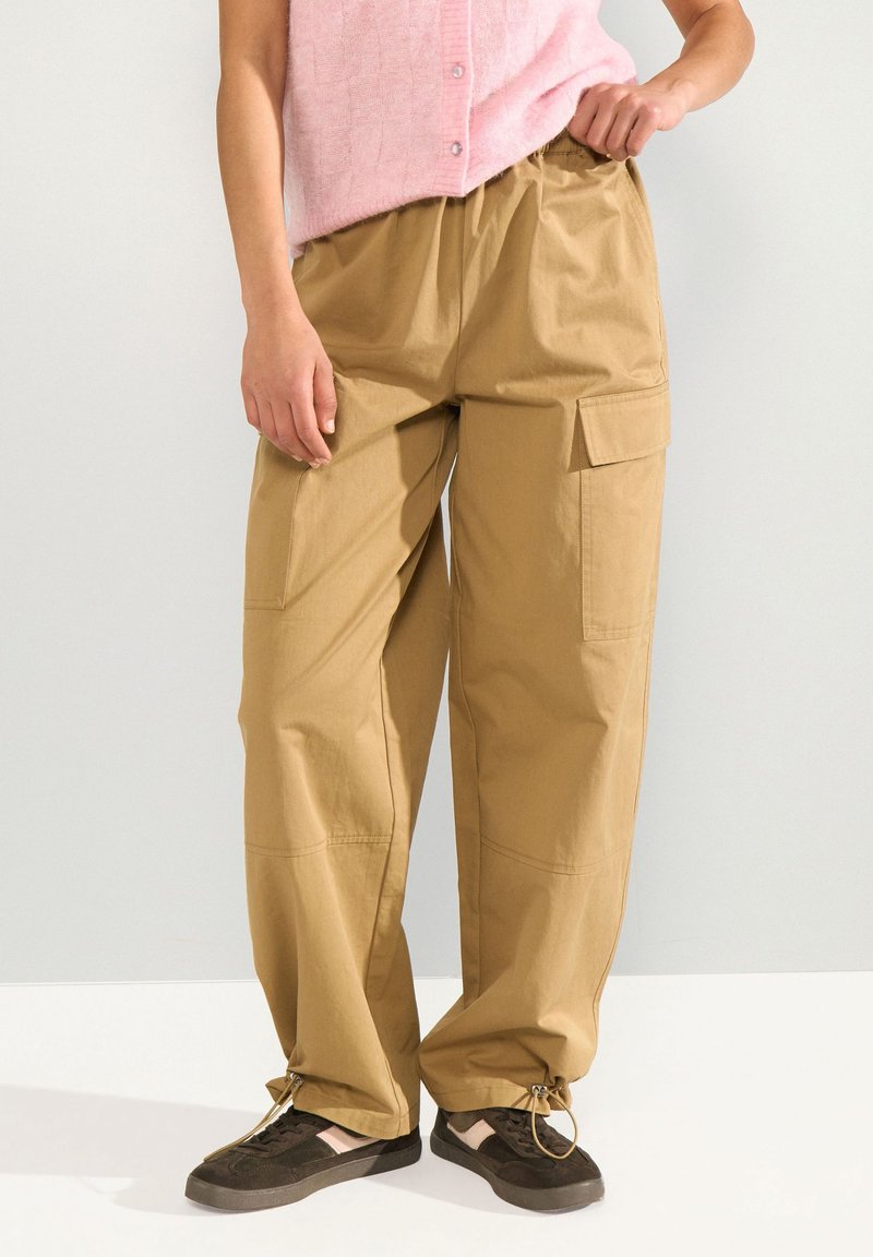 Personne portant un pantalon cargo ample beige avec des bas à cordon, des baskets noires et un haut rose sans manches, debout contre un mur uni.