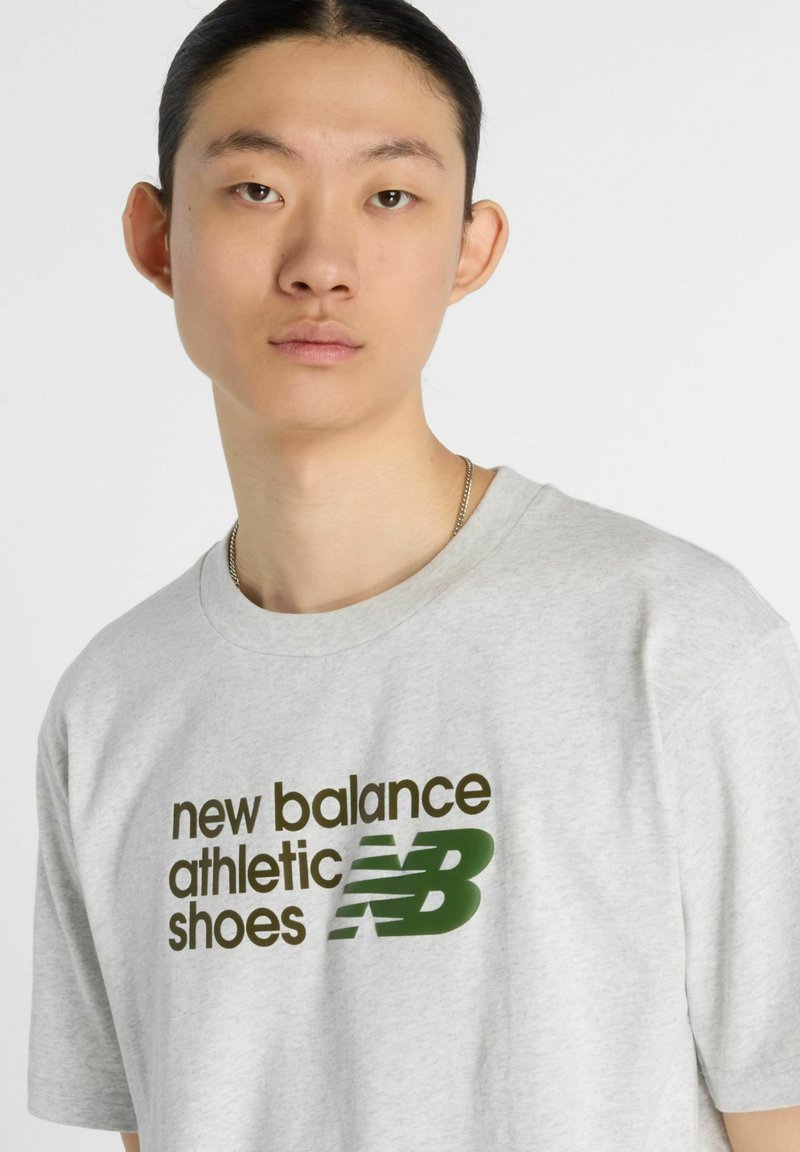 Grijze katoenen T-shirt met vetgedrukte zwarte en groene tekst "new balance athletic shoes" en een logo. Ronde hals en korte mouwen.