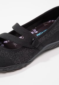 Zapato plano de mujer negra con dos correas ajustables, parte superior de tela texturizada, forro interior suave con patrón floral y suela flexible.