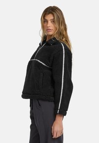 Schwarze Fleecejacke mit weißen Paspeln, Halb-Reißverschluss und Kängurutasche; zeichnet sich durch eine kastige Passform und ein strukturiertes, plüschiges Material aus.