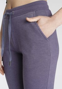 Lila Jogginghose mit geripptem Bund, Kordelzug mit Metallenden und seitlichen Taschen für Funktionalität. Weicher, strukturierter Stoff.