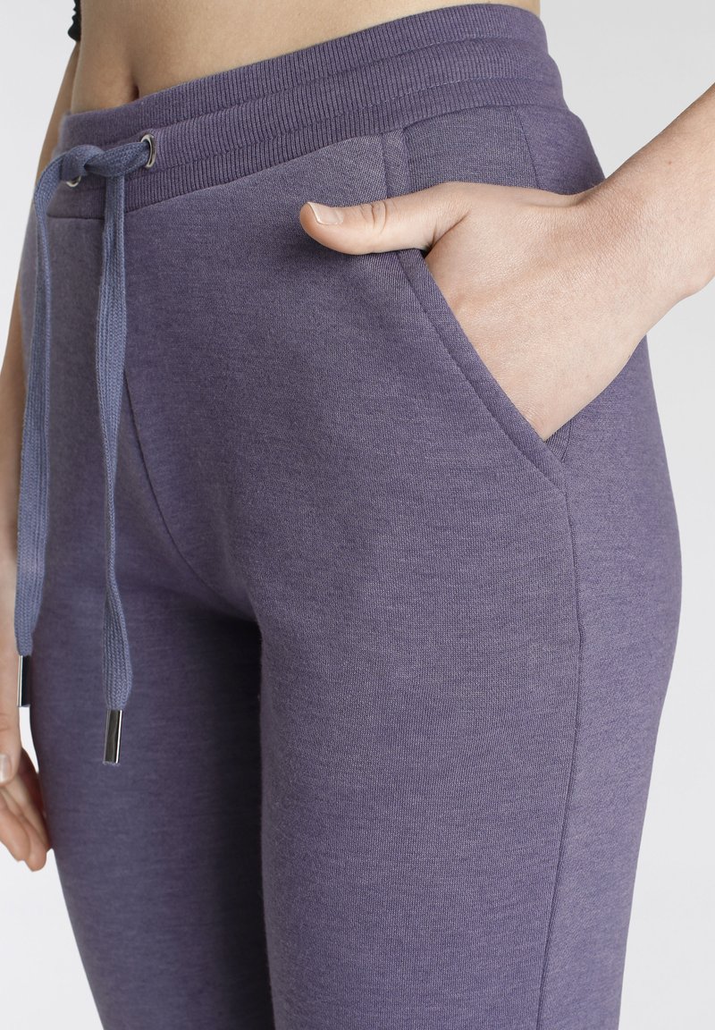Lila Jogginghose mit geripptem Bund, Kordelzug mit Metallenden und seitlichen Taschen für Funktionalität. Weicher, strukturierter Stoff.